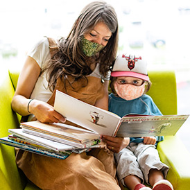 A woman and toddler sitting together, wearing masks and reading a picture book