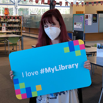 Library staff member Leah wearing a mask and holding a large sign with text on it reading: I love #MyLibrary