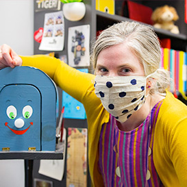A woman wearing a mask posing next to a blue mailbox that has a smiling face painted on the front