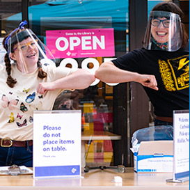 Two library employees wearing face shields smiling and posing outside at curbside pick-up