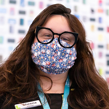 A woman wearing glasses and a face mask smiles at the camera. Her nametag reads Sheena.