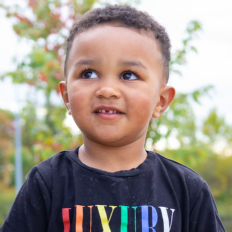 A preschool-aged boy standing outside with trees in the background, smiling and looking thoughtfully to the side.