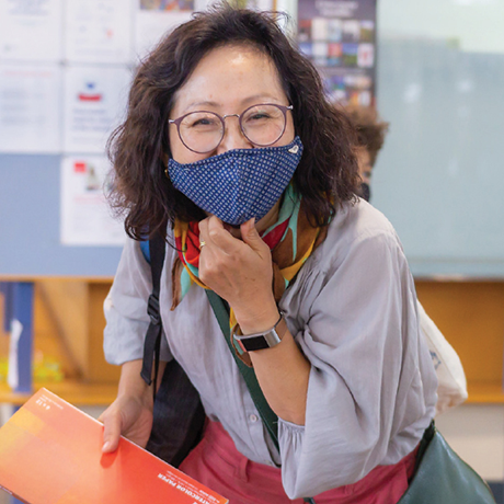 Library staff member with shoulder-length brown hair, wearing glasses and a mask, smiling welcomingly toward the camera