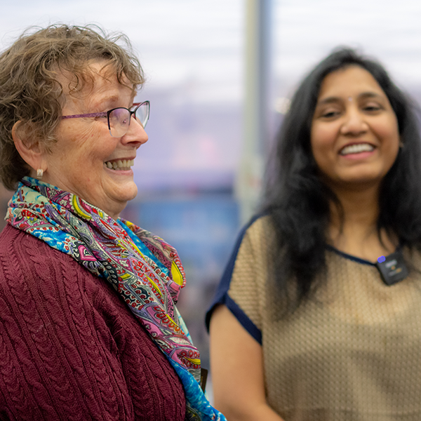 Two women, Carol and Siva, stand inside Alderney Gate Public Library, among book shelves, speaking warmly to one another.