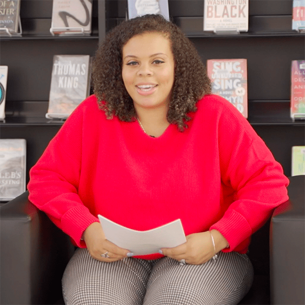 Rajean Willis sits, interviewer style, in a gray living room-style chair in front of a black bookshelf on the 5th floor of Central Library.