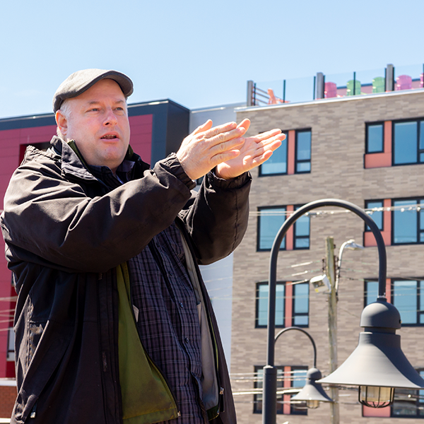Andrew Curran, IT staff member, stands on the roof of Halifax North Library on a sunny day, wearing a dark jacket and news cap, gesturing with his hands to show the reach of WiFi signals.