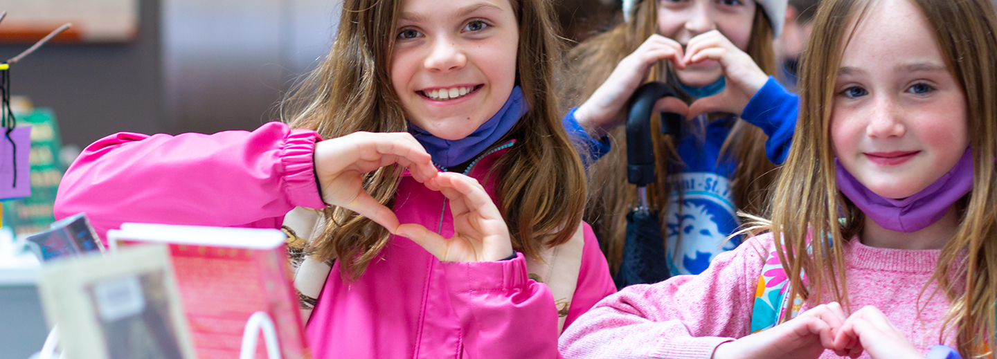 Three middle-grade age girls with mid-length straight brown hair, pose to camera, holding up their hands in heart formations. They are smiling broadly and all wearing bright pink and blue tones. The first floor of Central Library can be somewhat seen in the background.