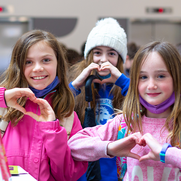 Three middle-grade age girls with mid-length straight brown hair, pose to camera, holding up their hands in heart formations. They are smiling broadly and all wearing bright pink and blue tones. The first floor of Central Library can be somewhat seen in the background.