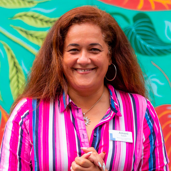 Amani stands outdoors in front of a colourful street mosaic, wearing a bright pink striped blouse and Library nametag. She has long brown hair and is smiling widely to camera.
