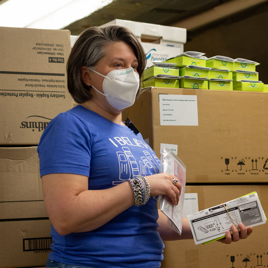 Tracey Stone, a Library Administration staff member with short brown hair, wearing a mask and blue t-shirt, stands in the basement of Alderney Gate Public Library, surrounded by rapid test kit boxes.